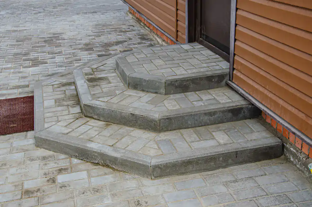 Three wide concrete steps with geometric patterns lead up to a dark door on a brown-sided building, with a paved walkway surrounding the steps.