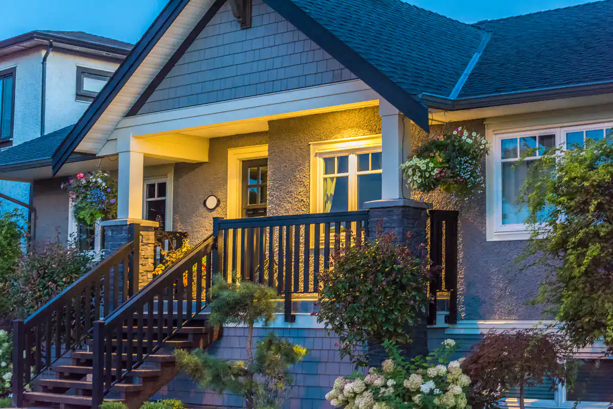 A cozy, well-lit house with a front porch, dark railings, hanging flower baskets, and landscaped shrubs and trees in the garden during dusk.