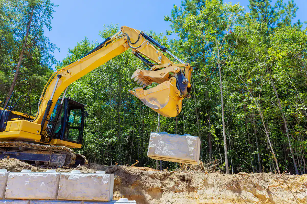 A yellow excavator lifts a large concrete block at a construction site surrounded by trees and dirt under a clear blue sky.