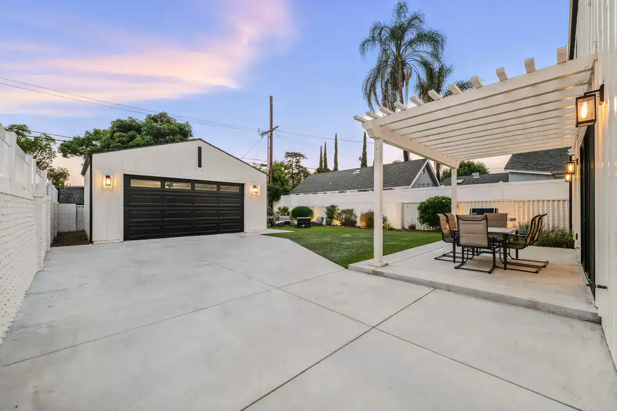 Modern backyard with a white pergola over an outdoor dining set, a manicured lawn, a white detached garage with black doors, and a paved driveway at sunset. Tall palm trees and a white fence are in the background.