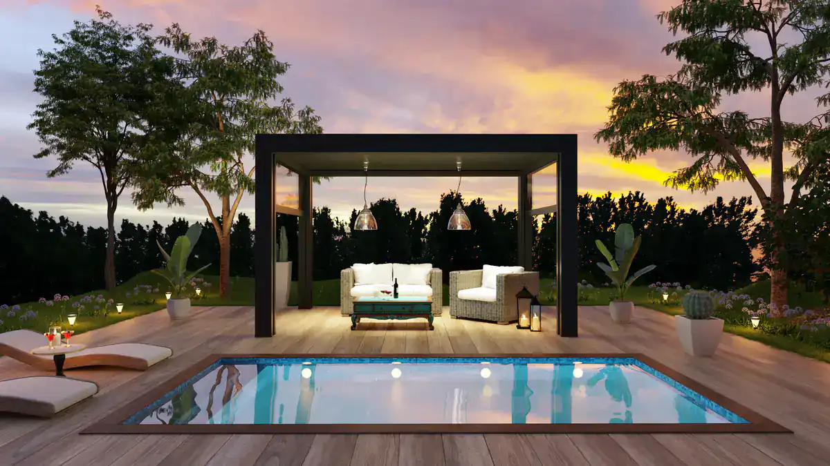 Modern backyard with a pool, poolside lounge chairs, a covered seating area with a sofa and armchairs, plants, and lanterns, set against a colorful sunset sky and surrounded by greenery.