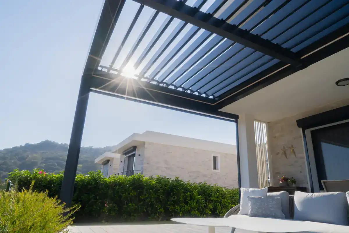A modern patio with a sleek pergola lets sunlight through its slats. In the background are green bushes, hills, and a contemporary house. The patio features a chair and small table with cushions.