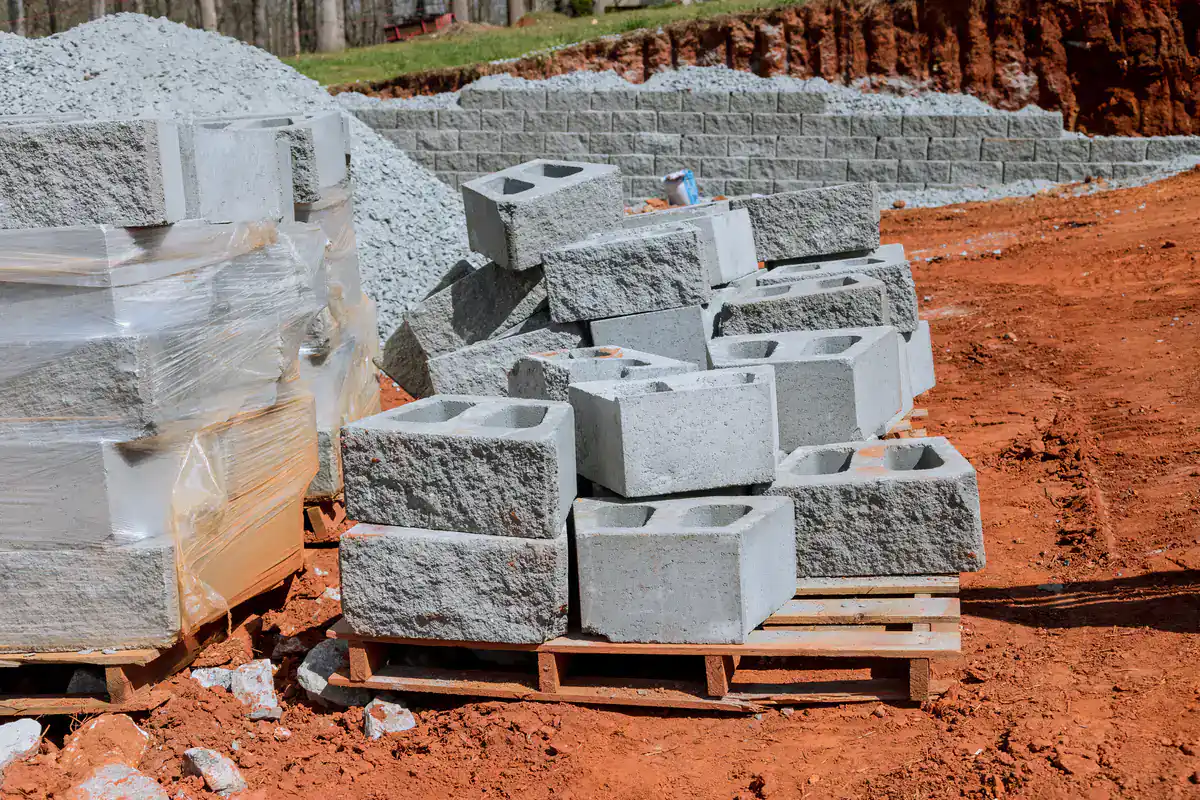 Stacked concrete cinder blocks sit on wooden pallets at a construction site with red dirt and gravel. Some blocks are neatly wrapped in plastic, while others are scattered or stacked loosely.