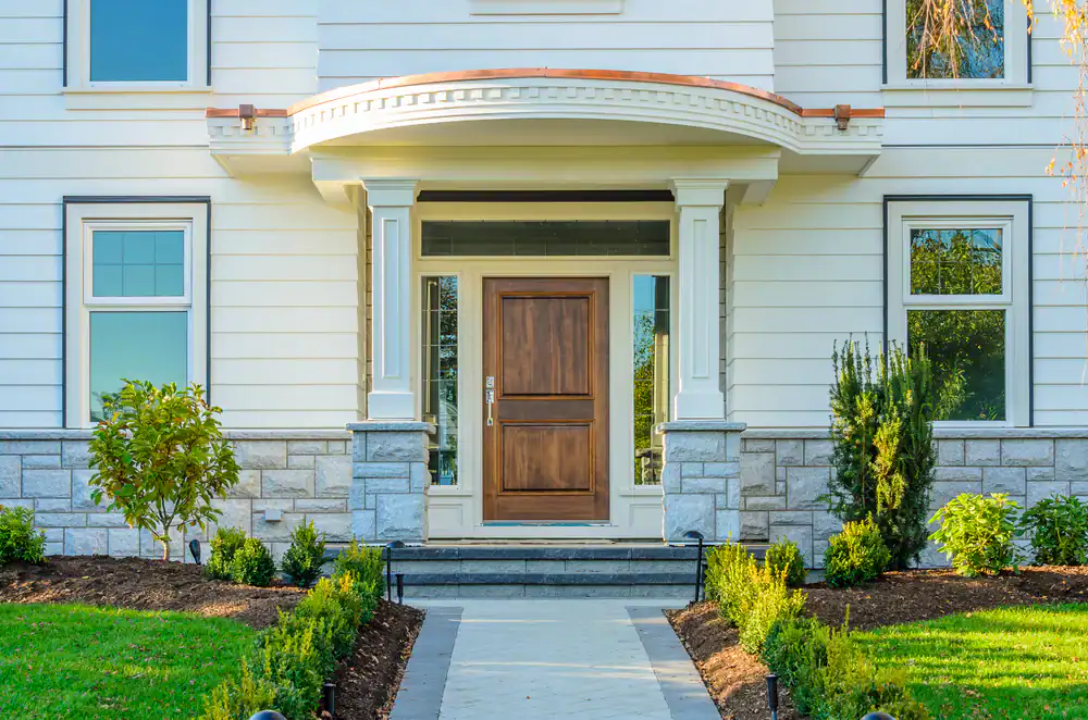 Front entrance of a house with a wooden door, white columns, stone accents, and symmetrical windows. Neatly trimmed bushes and plants line the walkway leading to the porch.