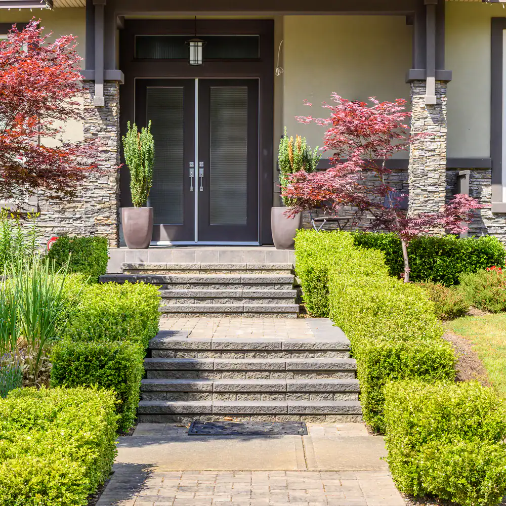 Front entrance of a house with double glass doors, stone pillars, potted plants, trimmed green hedges, a few red-leaved trees, and a short set of steps leading to the porch.