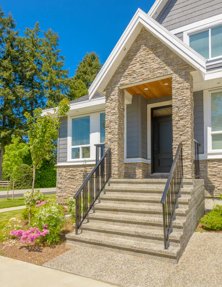 Front entrance of a modern house with gray siding and stone facade, wide concrete steps, black railings, a black front door, and landscaped garden with flowers and small trees under a clear blue sky.