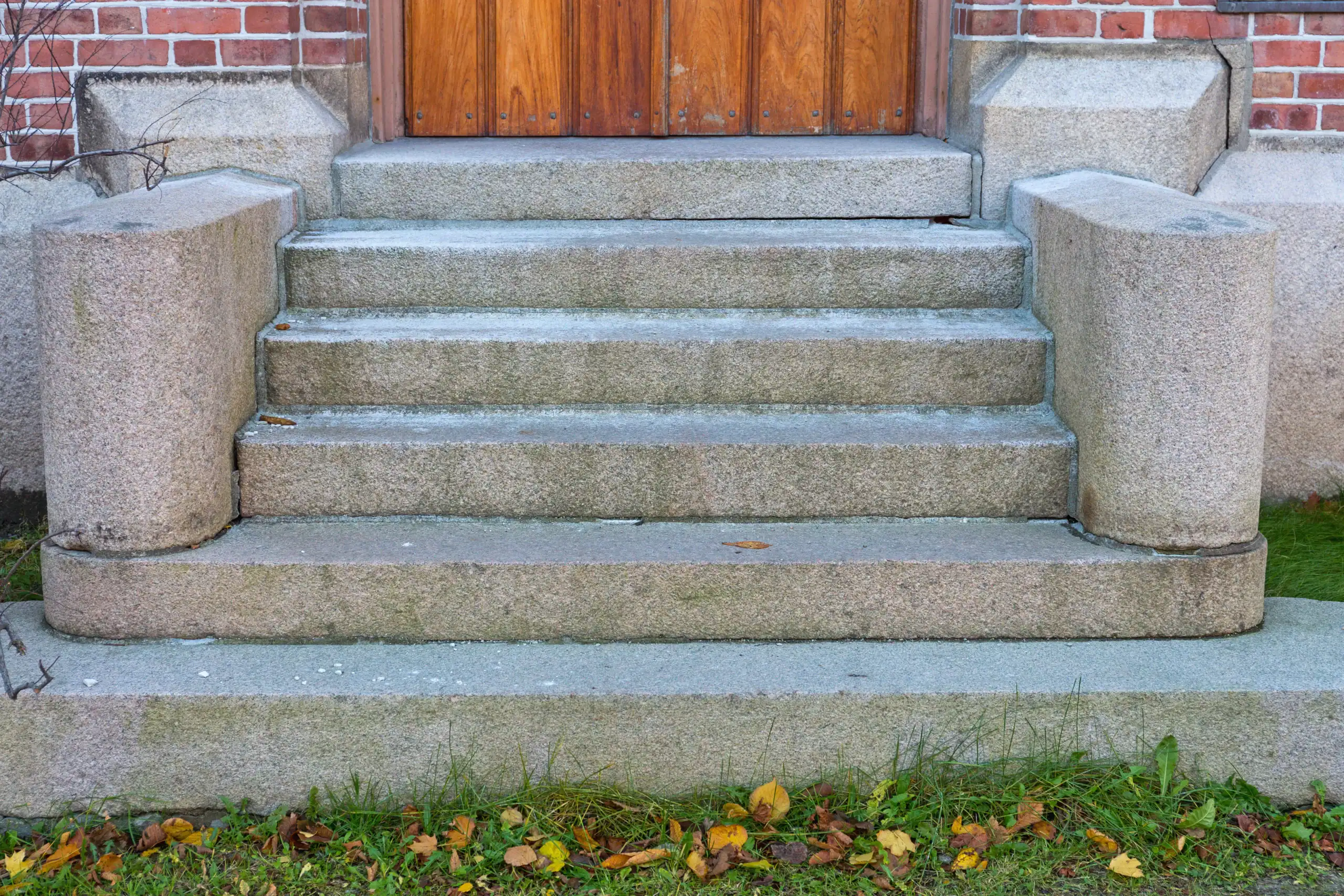 Concrete steps with rounded sides lead up to a wooden door. The steps are bordered by a small patch of grass and fallen autumn leaves, and the building's exterior wall is made of red brick and stone.