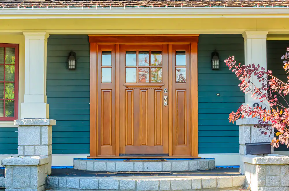 A wooden front door with decorative glass panels, flanked by two wall lanterns, set in a blue house with white trim and stone steps leading to the entrance.