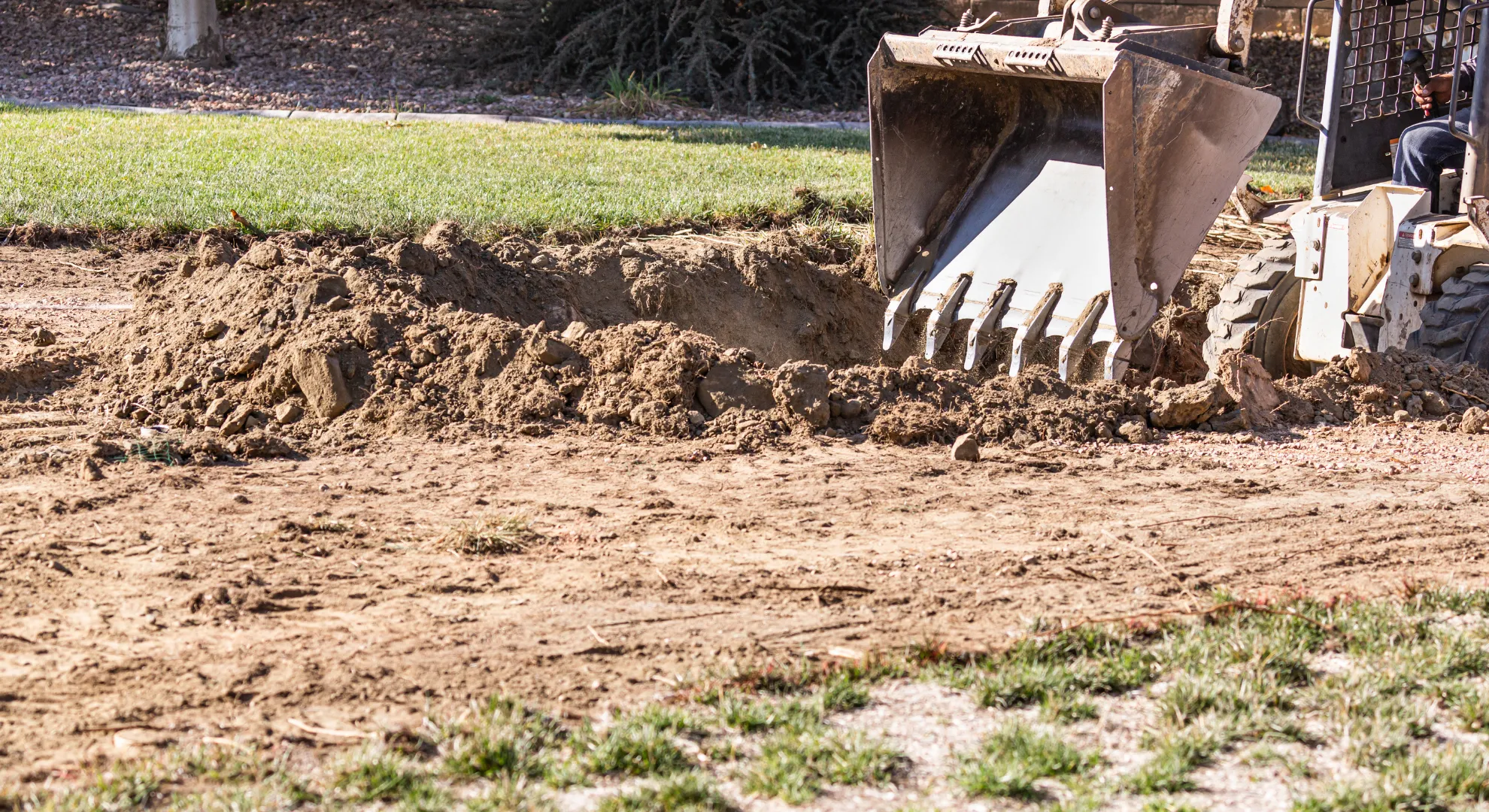 A construction vehicle with a bucket attachment digs in the dirt on patchy grass at a work site.